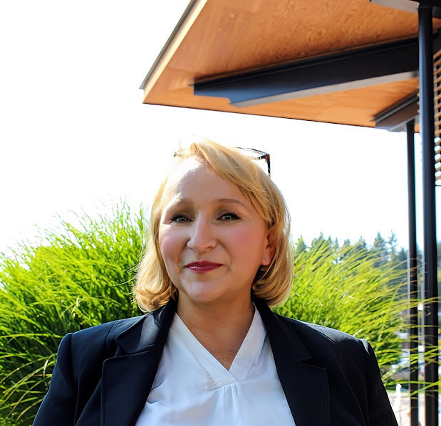 Picture of Corina Pfeil, she has sholder leangth blond hair, wearing a black blazer and white shirt, is smiling as the picture is taken outdoors in front of a community center.